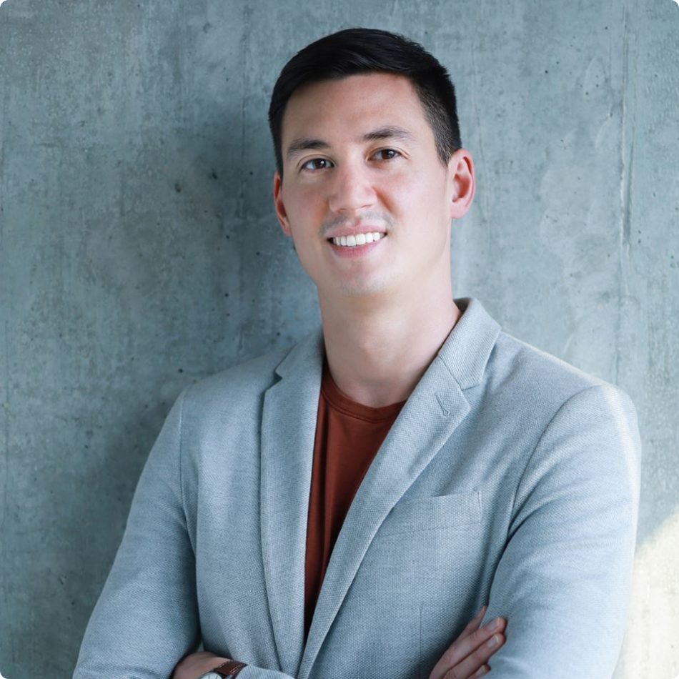 Mark McDowell standing with arms folded in a light grey suit against a bare concrete wall