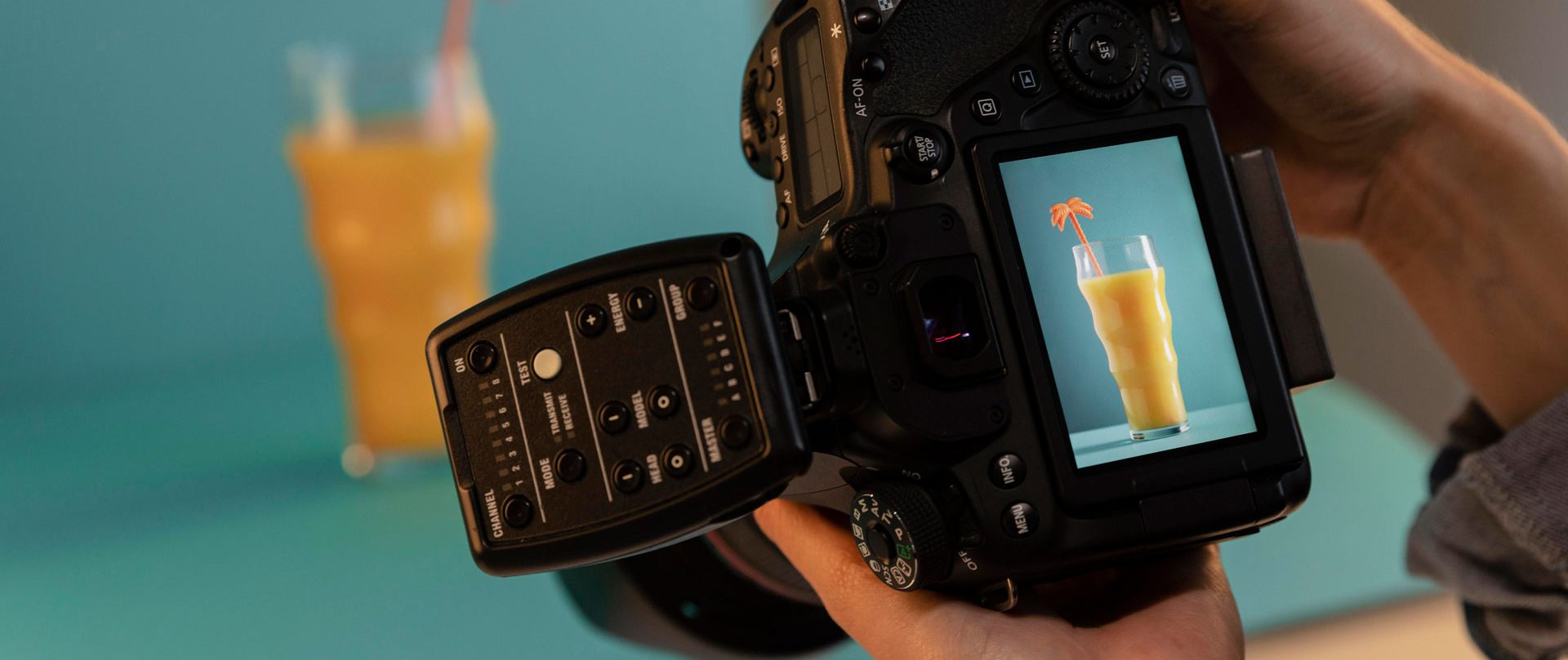 Hands holding a DSLR camera photographing a glass of orange juice on a table, the shot visible on the rear screen