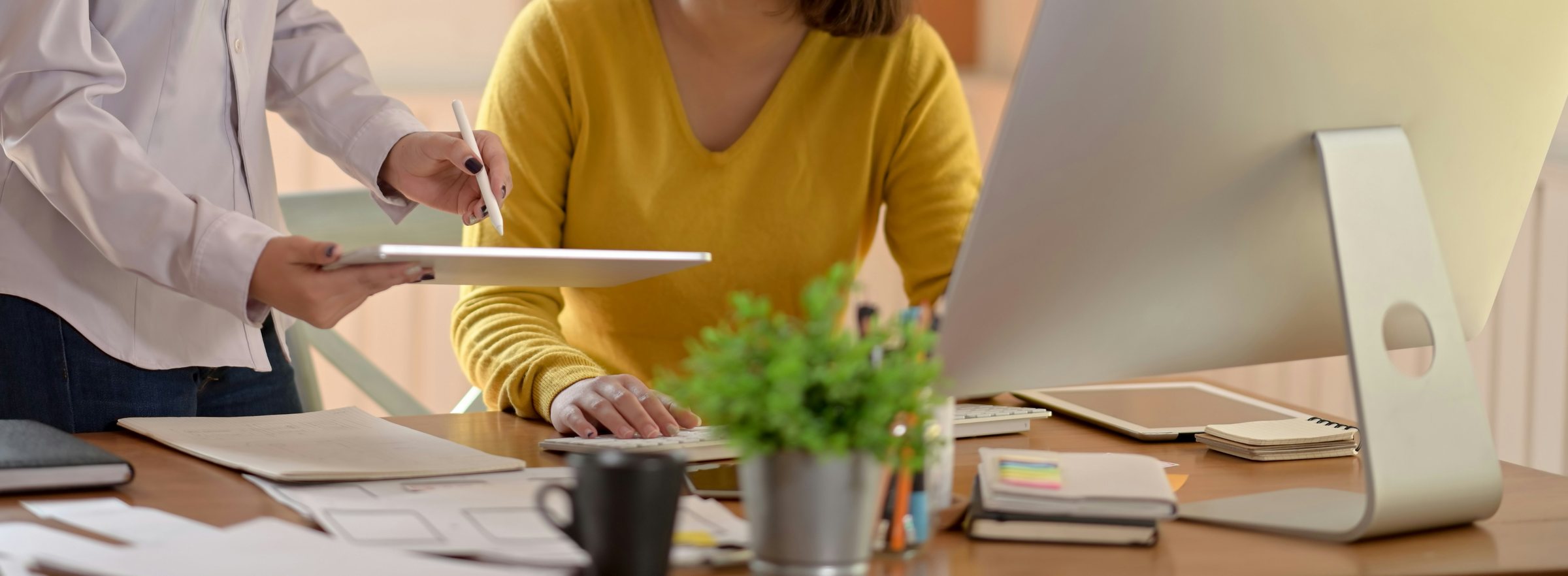 Colleague pointing a stylus at a tablet held over a desk while another works at a desktop computer